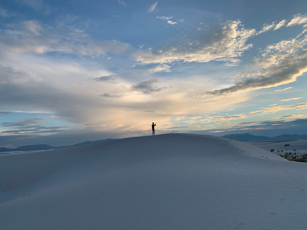 a person on a sand dune taking a picture at sunset in the west with mountains in the background