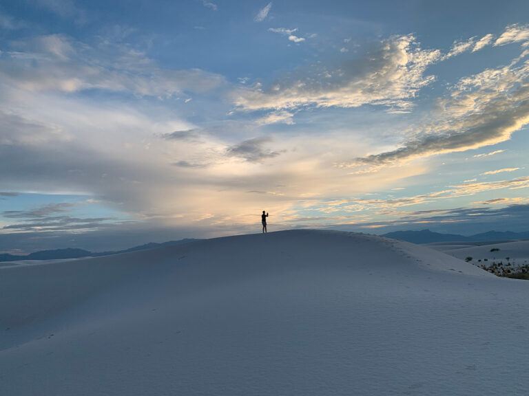 a person on a sand dune taking a picture at sunset in the west with mountains in the background