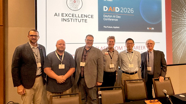 A group picture of panel participants smiling, standing on a stage in an auditorium.